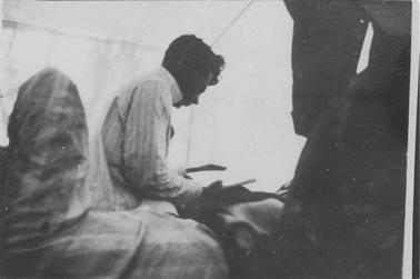Interior of tent. Photo taken from right side of young man wearing pajamas, seated on bunk reading a book.  Dark shape right forehand side.