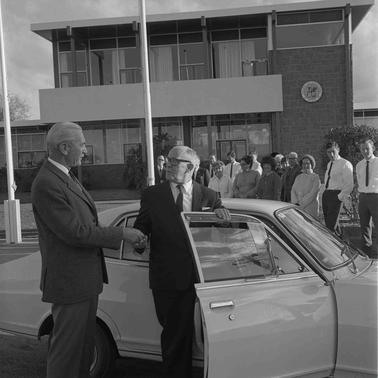 Two men at an open car door with a group of others looking on from the backgrouno date.