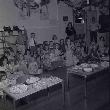 A group of primary school chidren dressed for a party sitting on the floor in rows near two tablkes laden with treats.