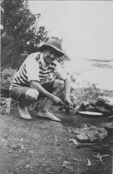 Camping scene. Young man squatting, holding a frying pan over camp fire, looking at camera. Wearing soft shoes, long shorts, shirt with sleeves rolled up, striped top and felt hat. Ocean and reef in right backgrouno date. Bushes in left backgrouno date.