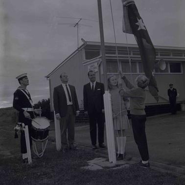 Near a school building a boy and girl are raising a flag while two men and a RAN drummer stand by.