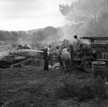 Six men and a fire truck at a fire scene.  One man is holding a running fire hose.