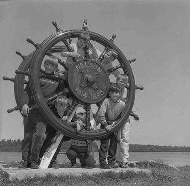 Seven boys posing at a ship’s wheel.