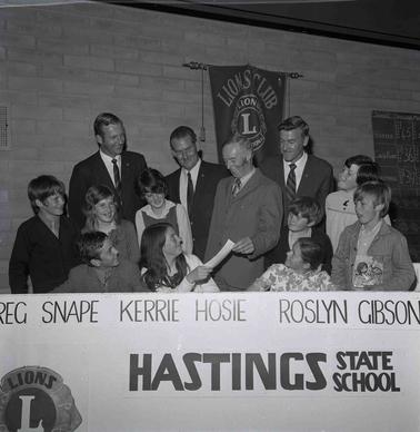 A group ofadults andchildren sitting and standing behind a banner with Hastings State School and other wrting on it.  One man, standing is handing a certificate to a woman, sitting,