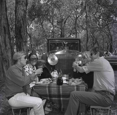Two couples picnicking under trees in front or a Rolls Rouyce car.