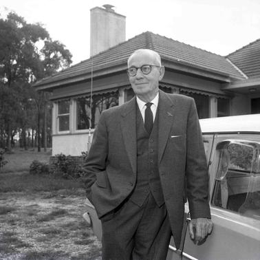 A suit wearing man standing by a car in front of a home.