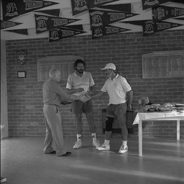 Three men, two shaking hands, in a clubhouse near a cloth covered table with plastic wrapped items on it.  Two honour rolls the wall are labelled ‘Somers Tennis Club’ and many pennants hang from the ceiling.