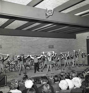 Eleven youn girls and one boy in performance costumes dancing on a stage in front of an audience.