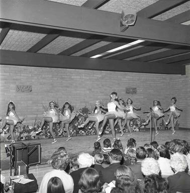 Eight young girls and one teenage girl dressed in tutus  performing a dance on a stage in front of an audience.