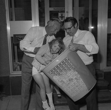 A girl reaching for a raffle ticket in a basket with the help of two men.