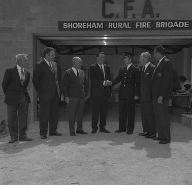 Seven men in suits, one in uniform,  in front of the Shoreham CFA building.  Two of the men are shaking hands.