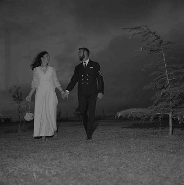 Woman in qa long pale frock and man in RAN uniform holding hands in an outdoor setting at night.  A young pine tree is bending with the wind on the right.