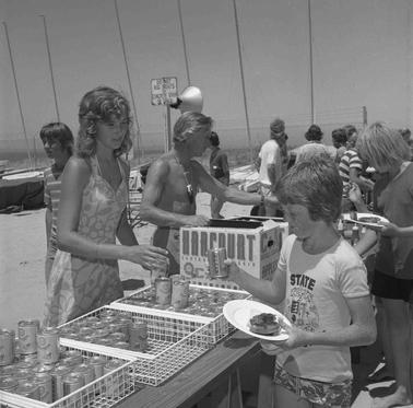 Beach scene with a food stall with children carrying barbeque food and collecting cans of soft drink.  Two girls serving out the provisions, other people are in the backgrouno date.