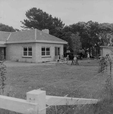 A pale brick cottage with five children playin on a lawn.  One child is walking along an eleveted board.