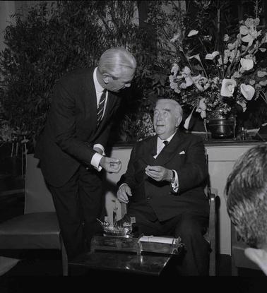 Two men in suits conversing in front of a floral arrangement, one is seated at a low table which has a model metal square rigged ship model on it.