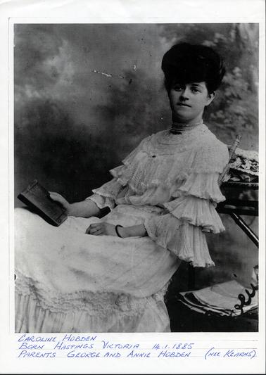 This image shows a young, beautifully dressed woman, reclining in a cane chair.  Her white dress has a high lace neckline and the sleeves have three tiers. Her black hair is piled high and she has three stands of pearls around her neck.  She is holding a book in her right hand.  