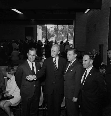 Four men in suits posing for photograph with people in the background and a view through a window to trees and parked cars.
