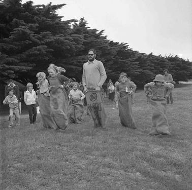 Four children and a man racing each other in a sack race with several children looking on.  Pine trees form the back drop.
