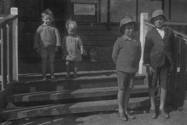 Three wooden stairs witha wooden floor beyono date.  Two boys are standing in front of the steps and infant boy and girl stand on top of the steps.  There is a bow labelled ‘Pure Velvet soap’ on the floor against the building and a poster ‘Promenade Estate - Balnarring East’ on wall.