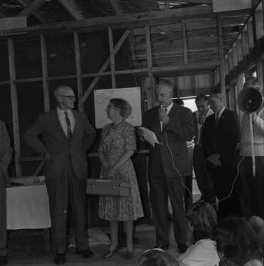 Six people on a dias in front of an audience inside an unfinished building, the wall frames are not clad.