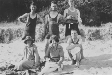 Six young adults on beach. Three men standing behind 2 seated women and one seated man. Men wearing one-piece bathing suits. One woman wearing hat and loose-fitting dress. Second woman with bobbed hair, wearing top and skirt. Women are seated on rug. Grasses and bushes in backgrouno date.