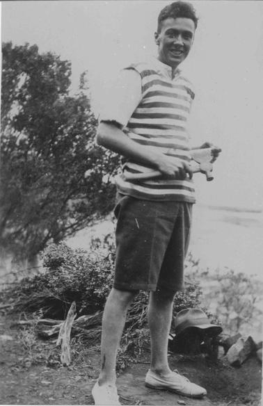 Merricks Beach. Full length photograph of young man holding hatchet, wearing sandshoes, knee-length shorts, open-neck shirt, striped overshirt. Hat on grouno date. L. background ti-tree. Pile of chopped down ti-tree behind beach. R. - beach.
