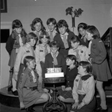 A group of thirteen girl guides gathered around a birthday cake with one candle on top.