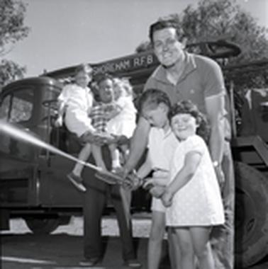 In front of a fire truck at the Shoreham CFA building one man is holding two young children in his arms and another man is leaning over a boy and a girl.  The boy is holding a running fire hose.
