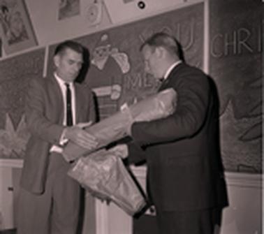 Two men exchanging parcels.  A blackboard is in the backgrouno date.