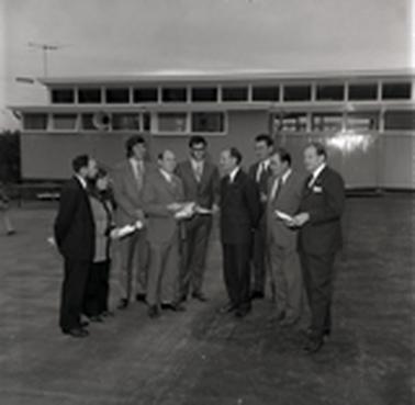Eight men in suits and one woman posing for a photograph in front of a school building.