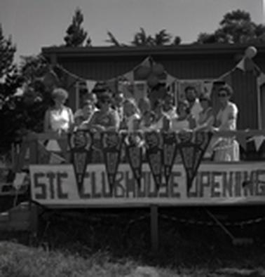 Fourteen men, women and children standing at a verandah rail festooned with pannants and a banner provlaiming STC Clubhouse Opening,
