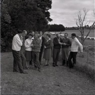 Eleven people (two are women) dressed in warm  outdoor clothing, are standing about in two groups near a paddock fence.  Beyond the fence lies a row of large pipes.  They seem to be discussing some issue.