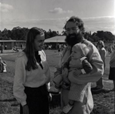 A couple (the man has a dark beard) with an infant at a sportsgrouno date.