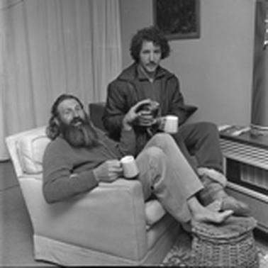 Two men dressed in warm casual clothes, one has his shoes off, sitting near a room heater.  they are relaxing with mugs of hot beverage.  The older, bearded man is in an armchair, the other is sitting on a chair holding up a yacht rope reel.