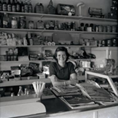 A woman standing behind a shop counter with shelves of goods behind her and magqazines on the counter.