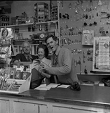 A woman and a man behind a store counter with shelves of goods behind them.