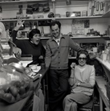 A man and two women either side of him, one woman is seated.  There are shelves of store goods behind them and a counter on the left.