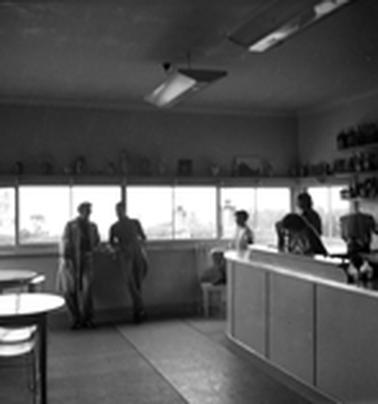 As store interior showing a counter with two women behind it, tables and chairs, two men chatting and a boy.  A row of windows on the far wall. 