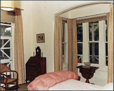Interior view of a bedroom with cream walls and gold curtains. Bay window to the right and another floor length window to the left. Bed in the foreground with a pink coloured duvet. Two pieces of dark wooden furniture and a cane chair to the left.