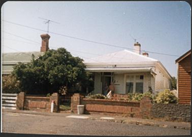 A white weather board residence with a iron pitched roof two chimneys with red brick detail a verandah
and a brick fence 