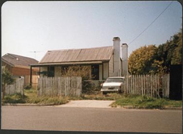 A weather board cottage with an iron roof, a verandah and two chimneys on right side of the building
a picket fence in the front of the cottage divided by gates