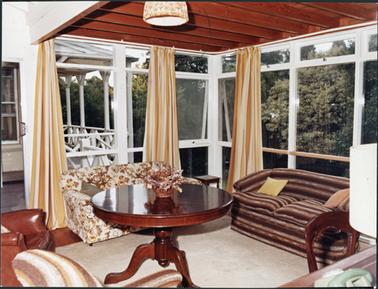 The sun room at Southdean, furnished with a round mahogany table, two couches against the windows and chairs just visible at the edge of the image. Striped yellow and cream curtains adorn the windows