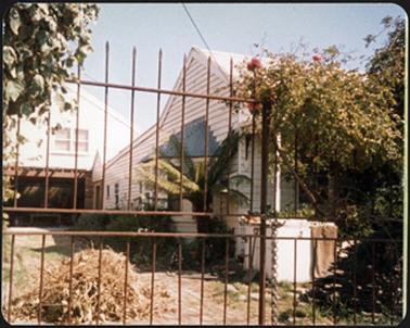 A side view of a white weatherboard residence with sloping roof built on at the back of the building
iron paling fence in the front of building with a barn in the background 