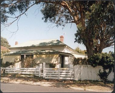 This image is of a white weather board residence with a green slanting iron roof and an enclosed verandah with two red brick chimneys, there is a white wooden fence in the front of the building and to the left of the front garden a small part has been divided off by a fence.