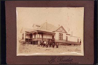 This is a weather board house, horizontal weather boards along bottom to window sill level on stilts, with steps leading to the garden there is “folk work” on gable, two chimneys and an attic on the left of the building there are nine male figures standing or kneeling at the front of the house
 