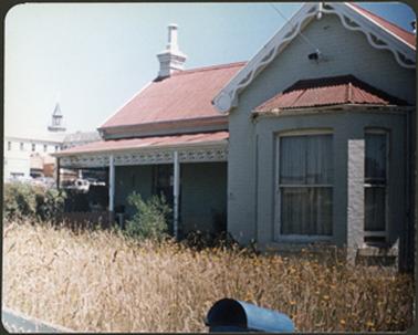 A front view of the house which is white brick with a red iron roof the verandah, double fronted with bay window