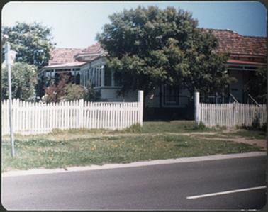 A white weatherboard residence with verandah and a red sloping roof in the front of the building a
white picket fence
