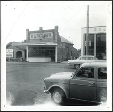The image depicts a small house to the left of the butchers shop with an arched porch and low brick fence. It has a ‘For Sale’ notice out the front. The Butchers Shop has a verandah with a lane way to the right and a more modern building which is a bank further to the right [E.S. & A. Bank] . There are two parked cars in the street.