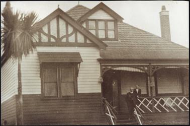 This is a weather board house horizontal weather boards along bottom to window sill level “Folk work” on gable, with two chimneys and an attic on the left of the building there is a verandah to the right with steps leading down to the garden there is one person standing on the steps and another person behind on the verandah