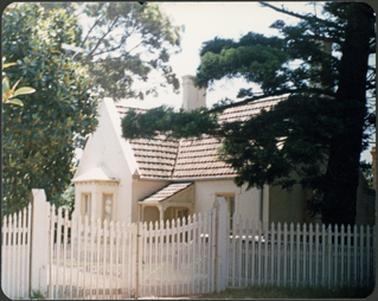 This house is of a side/front view, made of stucco brick with a tiled roof, bay window and small porch pine tree at right hand side of the house with picket fence and gate